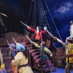 Captain Hook menacingly points his sword at Peter Pan as his pirate crew brandishes their swords in a theatrical scene from Peter Pan.