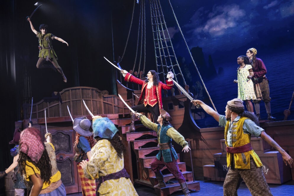 Captain Hook menacingly points his sword at Peter Pan as his pirate crew brandishes their swords in a theatrical scene from Peter Pan.