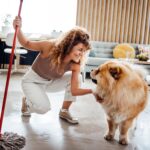 A woman happily mops her living room floor while her golden retriever dog sits and watches, showcasing a safe cleaning routine around pets.