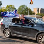 A man holds a Trump 2024 sign out the passenger window of a moving car.