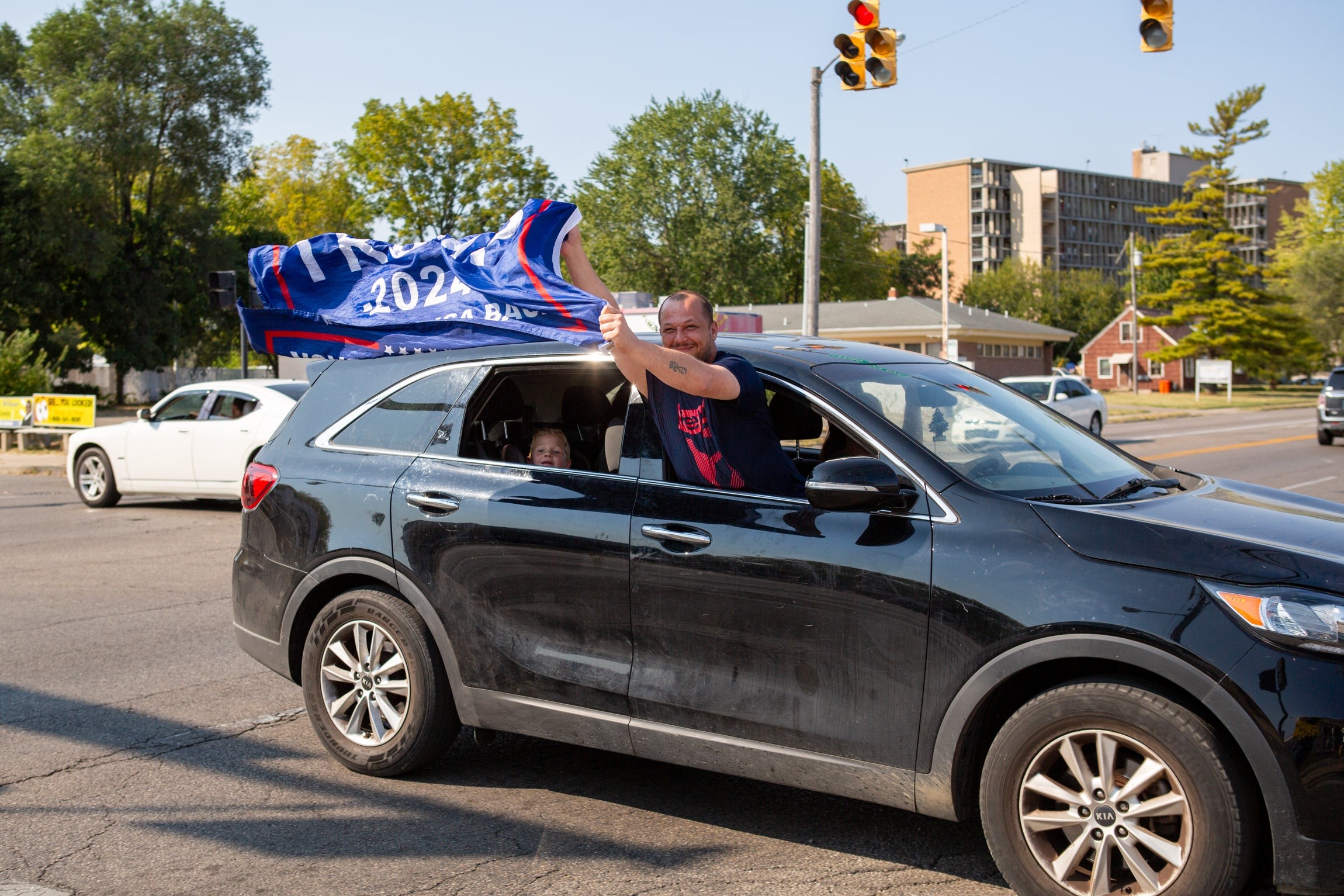 A man holds a Trump 2024 sign out the passenger window of a moving car.