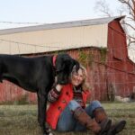 Great Dane standing next to a middle-aged woman outdoors on a farm.