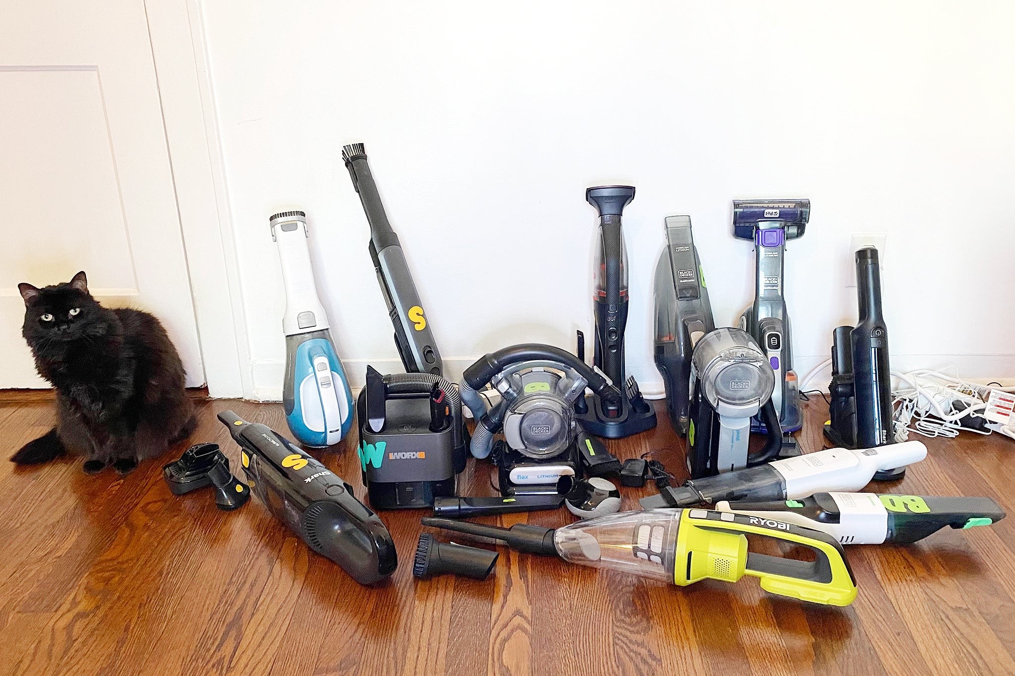 An assortment of handheld vacuums designed for pet hair removal are displayed on a wooden floor next to a black cat, showcasing various models and attachments.