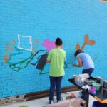 Two individuals collaboratively painting a vibrant mural on a blue wall, showcasing community art and creative expression.