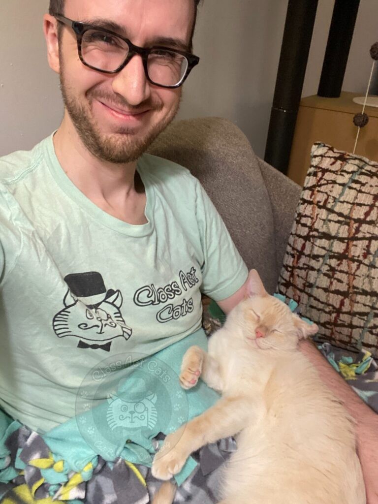 Joey smiling sitting on a couch with Prozac the cat curled up in his arm.